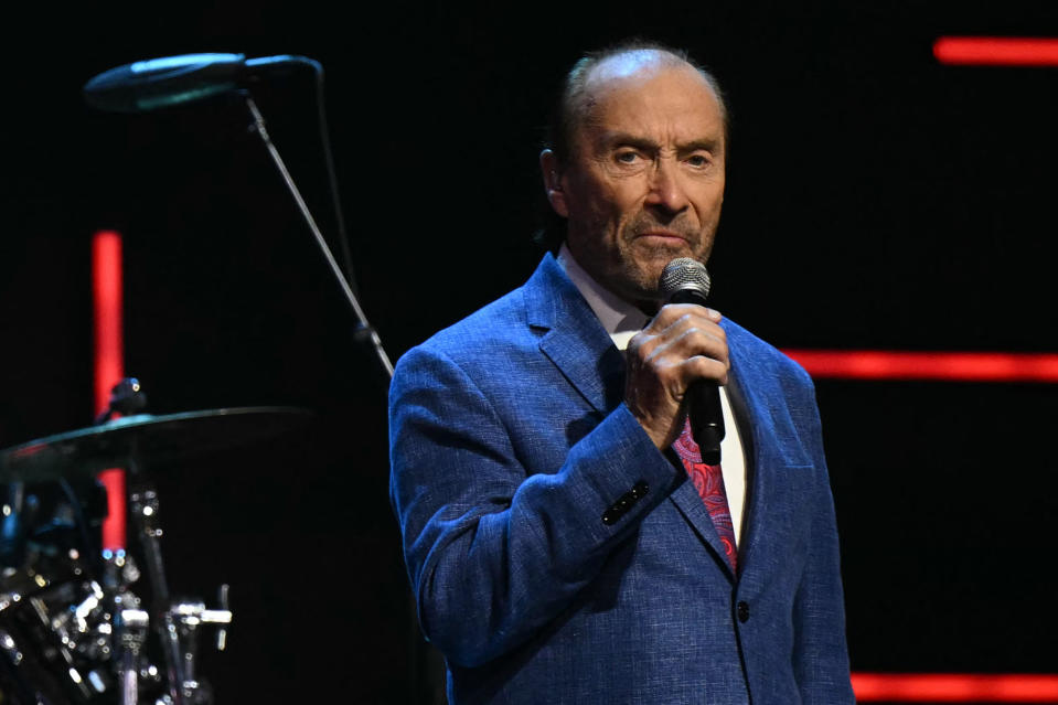 Lee Greenwood wearing a blue suit jacket with a pink tie, holding a microphone, performs on stage with a drum set partially visible behind him