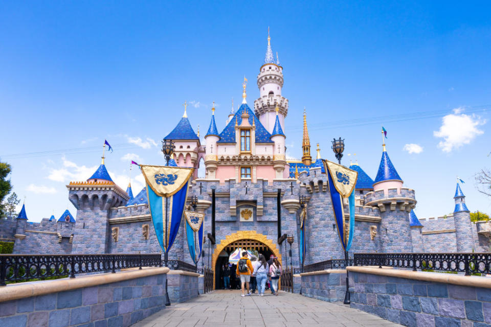 People walking through the entrance of Sleeping Beauty Castle at Disneyland. The castle features spires and flags