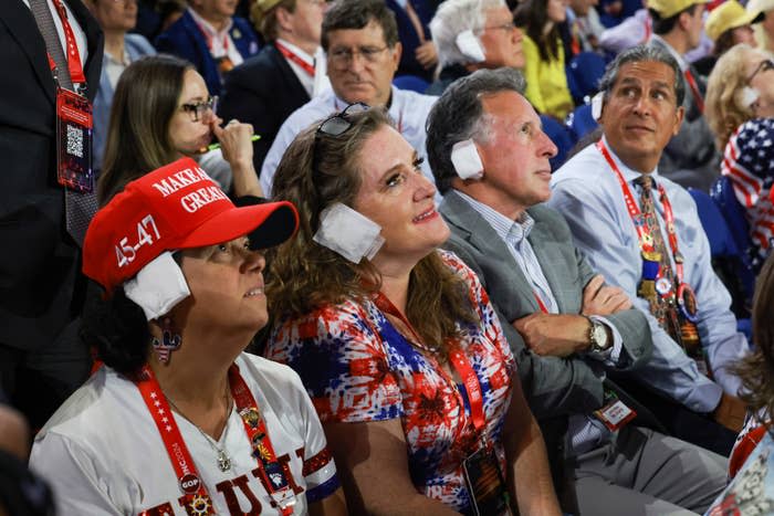 People at a Donald Trump rally, some wearing ear protection and patriotic clothing, sitting and listening attentively