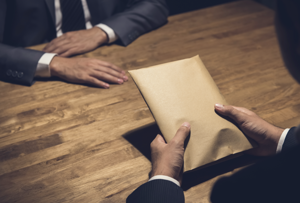 Two people in business attire sit at a wooden table while one person hands an envelope to the other