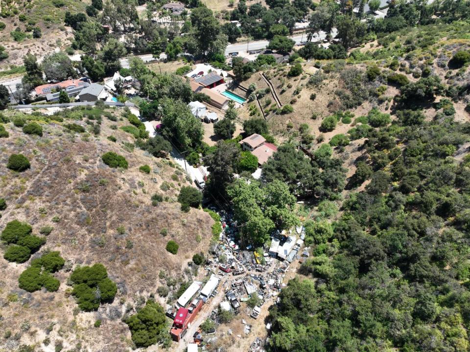 An aerial view of a rural property flanked by trees and grassy hills and crowded with vehicles.