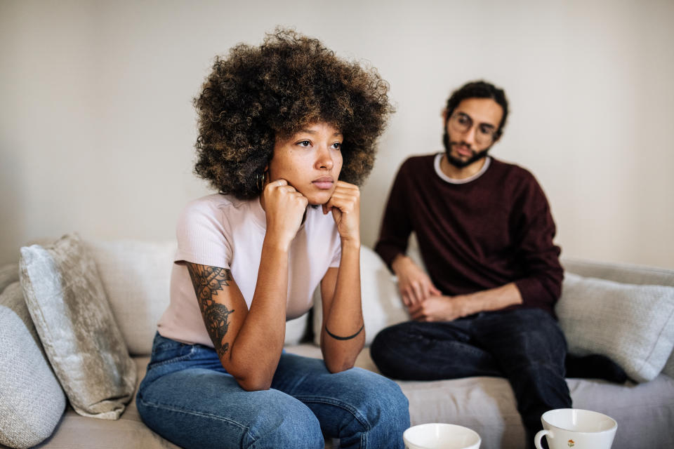 Woman with a thoughtful expression sits on a couch, man in the background looks at her. Both appear to be in a serious conversation