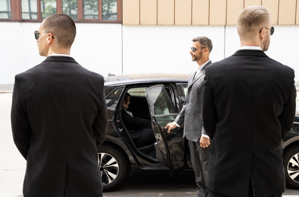 Three security personnel assist an individual exiting a car. The people are dressed in black suits. The context suggests an important or high-profile work-related event