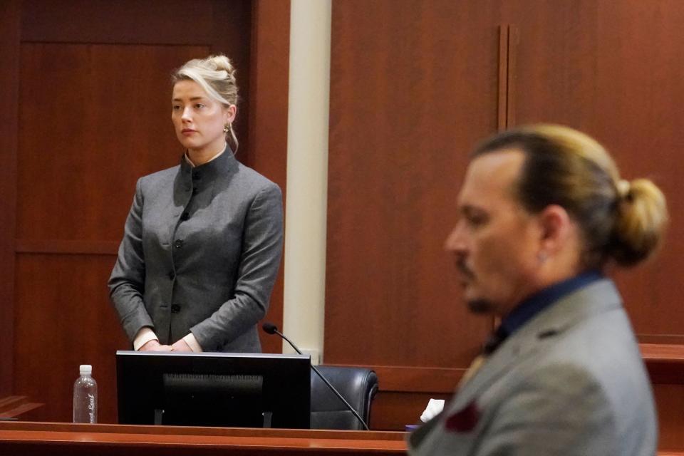 Johnny Depp and Amber Heard in a courtroom, both in gray suits. Amber stands near the bench, and Johnny sits at the table