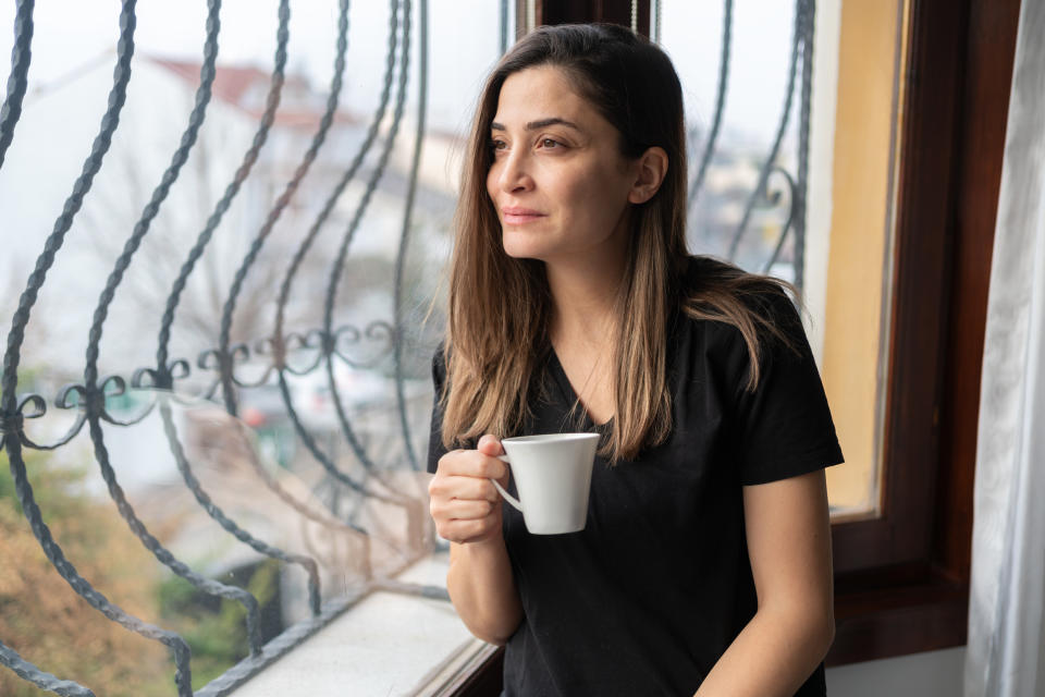 A woman in a casual t-shirt holds a coffee cup while looking thoughtfully out a window with decorative iron bars