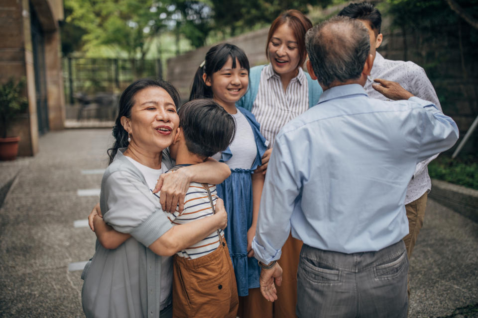 Group of six people, including grandparents, parents, and children, in an outdoor setting, smiling and hugging each other in a warm family moment
