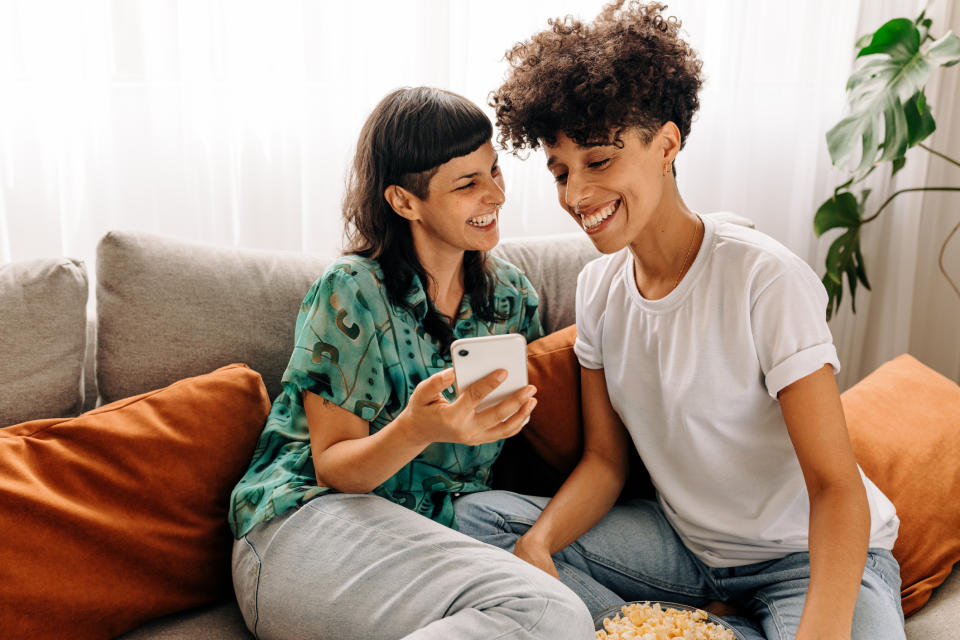 Two people sit on a couch smiling while one shows something on a phone screen to the other