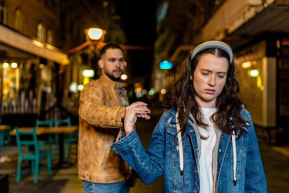 A man and a woman outside at night. The man is reaching for the woman's hand, while the woman looks upset and pulls away