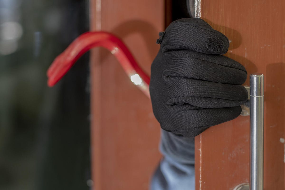 A gloved hand opens a red door with a crowbar at the ready in the background.