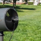 A black fan with misty water spraying from the front in a grassy backyard