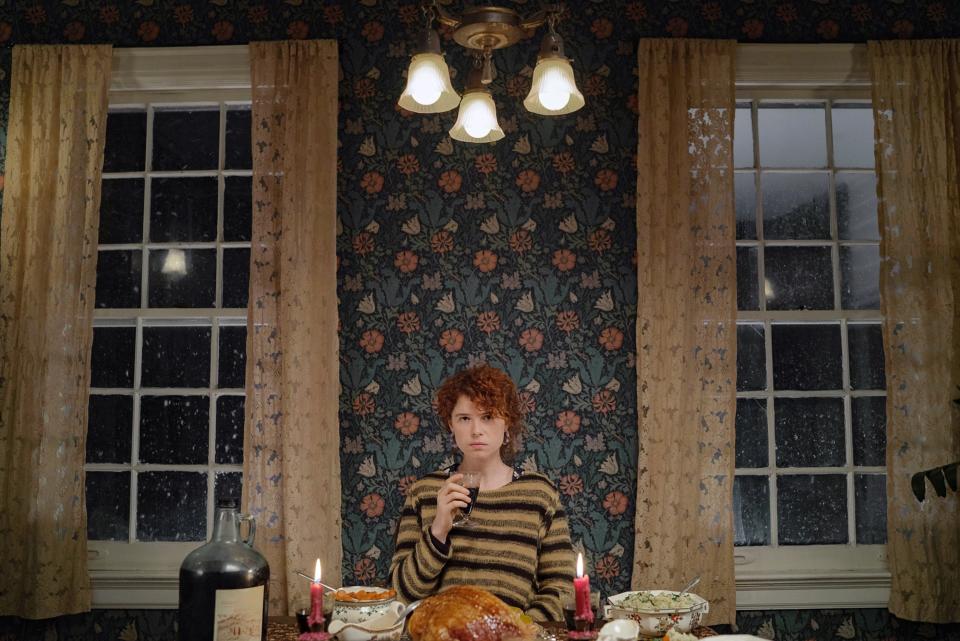 A person with curly hair, wearing a striped sweater, sits at a dining table with a roast turkey and candles in front of them in a vintage-styled room
