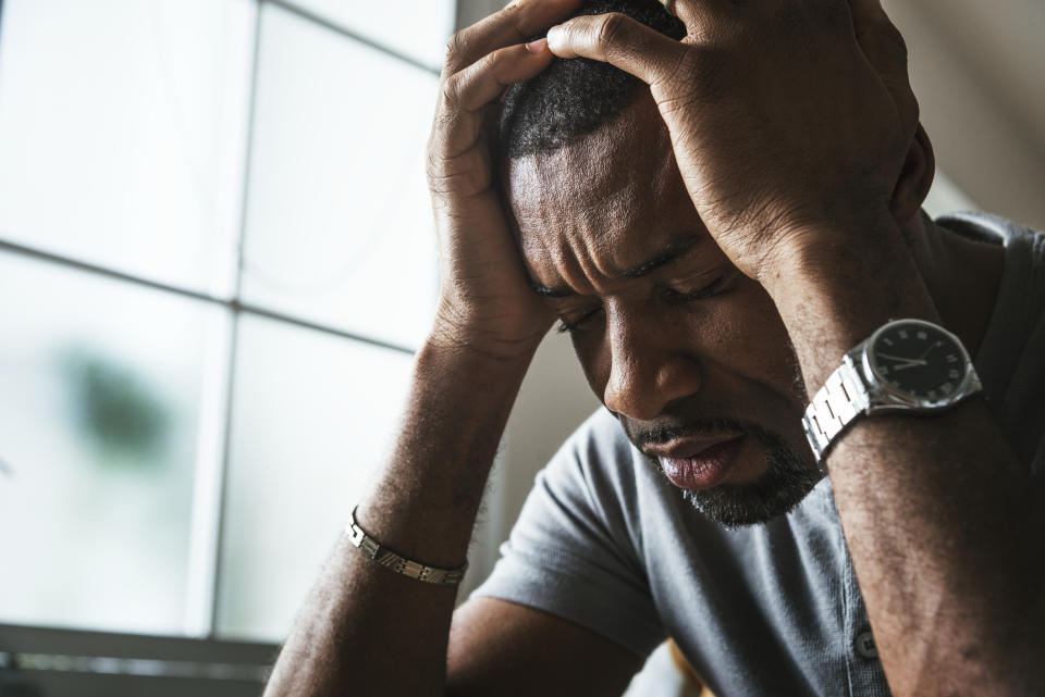 A man with a wristwatch and bracelet holding his head in distress near a sunlit window