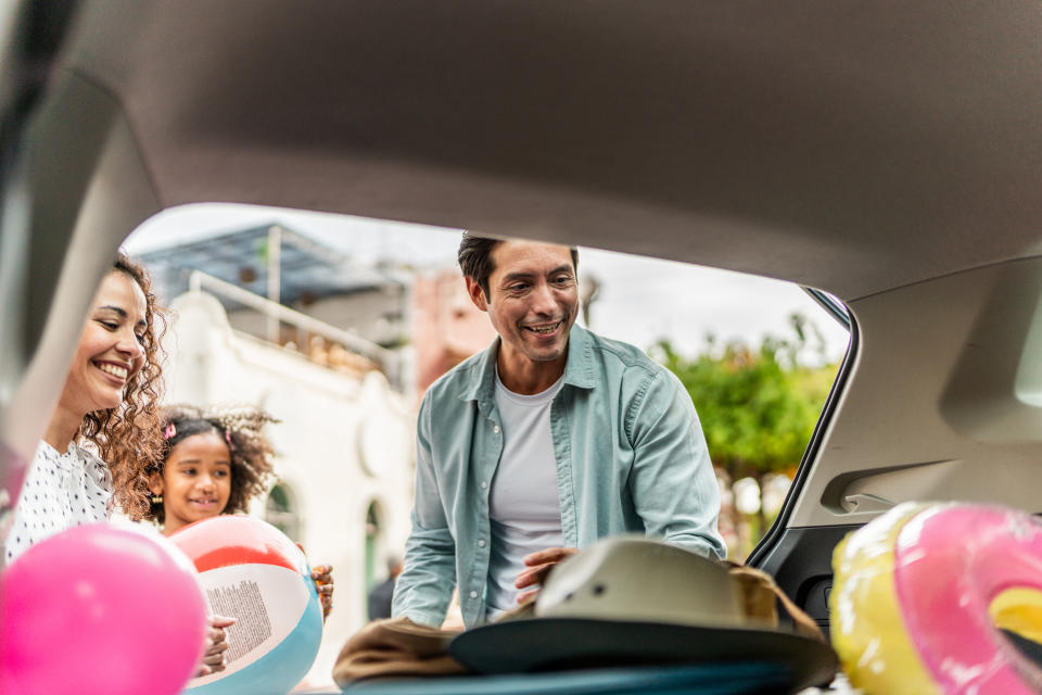 Family of three, including a man, a woman, and a young girl, happily packing items into a car trunk, possibly preparing for a beach trip