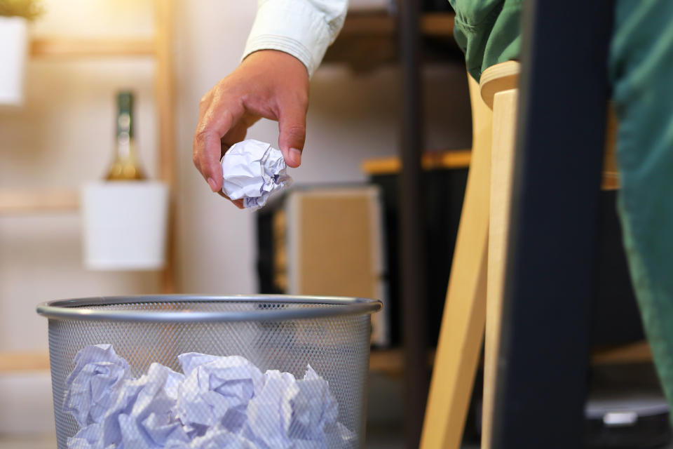 A hand drops a crumpled paper ball into a trash can filled with other crumpled papers in an office setting