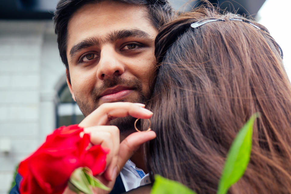 A smiling man hugs his fiancé and holds a ring