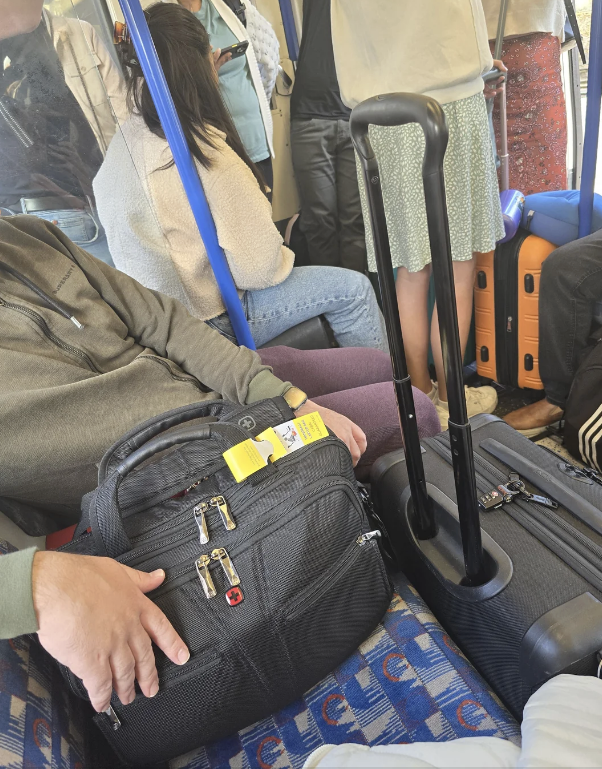 People seated and standing on a crowded train, some with luggage. Orange suitcase and black backpack are visible prominently. Faces are not clear