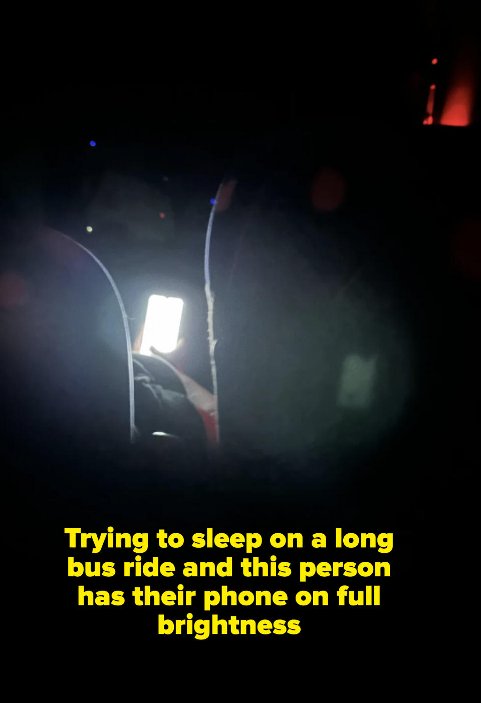A person holding a glowing phone in a dark setting, partially obscured by two seats in front of them