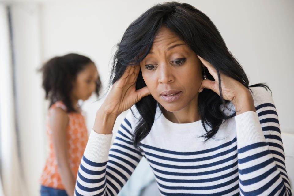 A woman in a striped sweater looks stressed, holding her temples, with a child in the background wearing a patterned dress