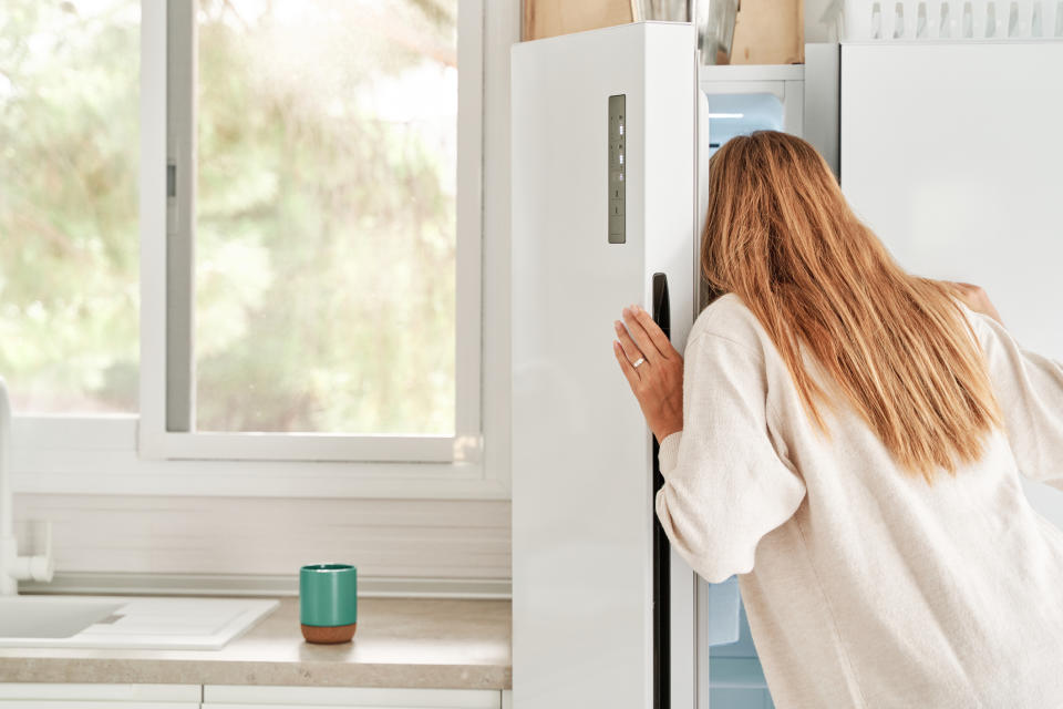 A person with light brown hair wearing a light sweater looks inside a refrigerator in a kitchen with a window in the background
