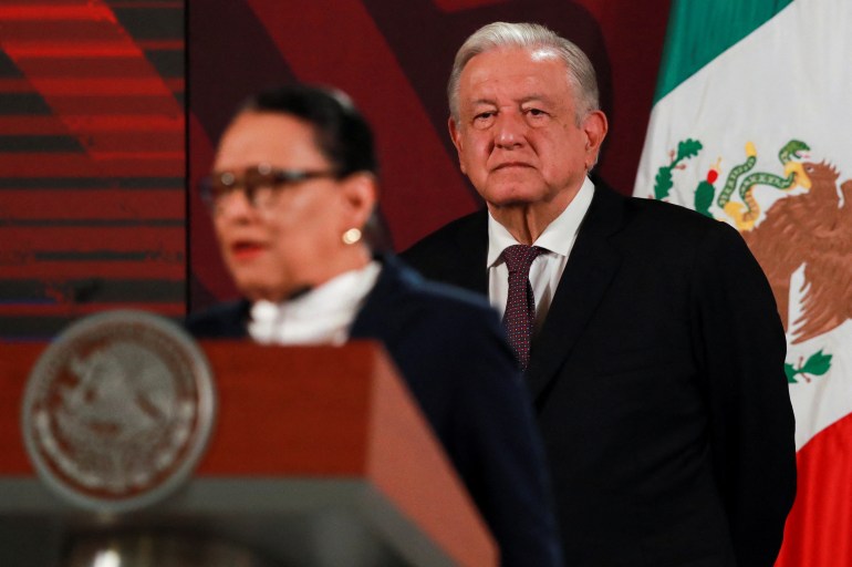Mexico's Security Minister Rosa Icela Rodriguez speaks at an official podium, while Andres Manuel Lopez Obrador and a Mexican flag stand behind her.