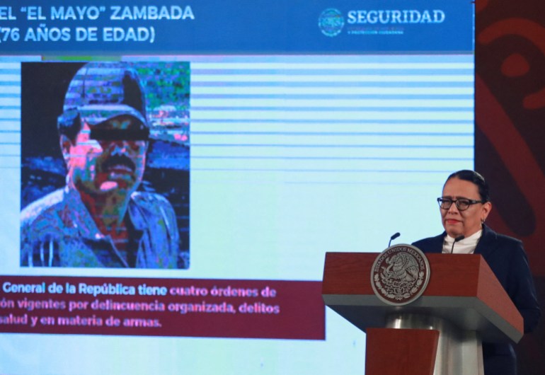 Rosa Icela Rodriguez speaks behind a government podium. Behind her is a screen with a slide show presentation, showing the drug lord El Mayo's face.