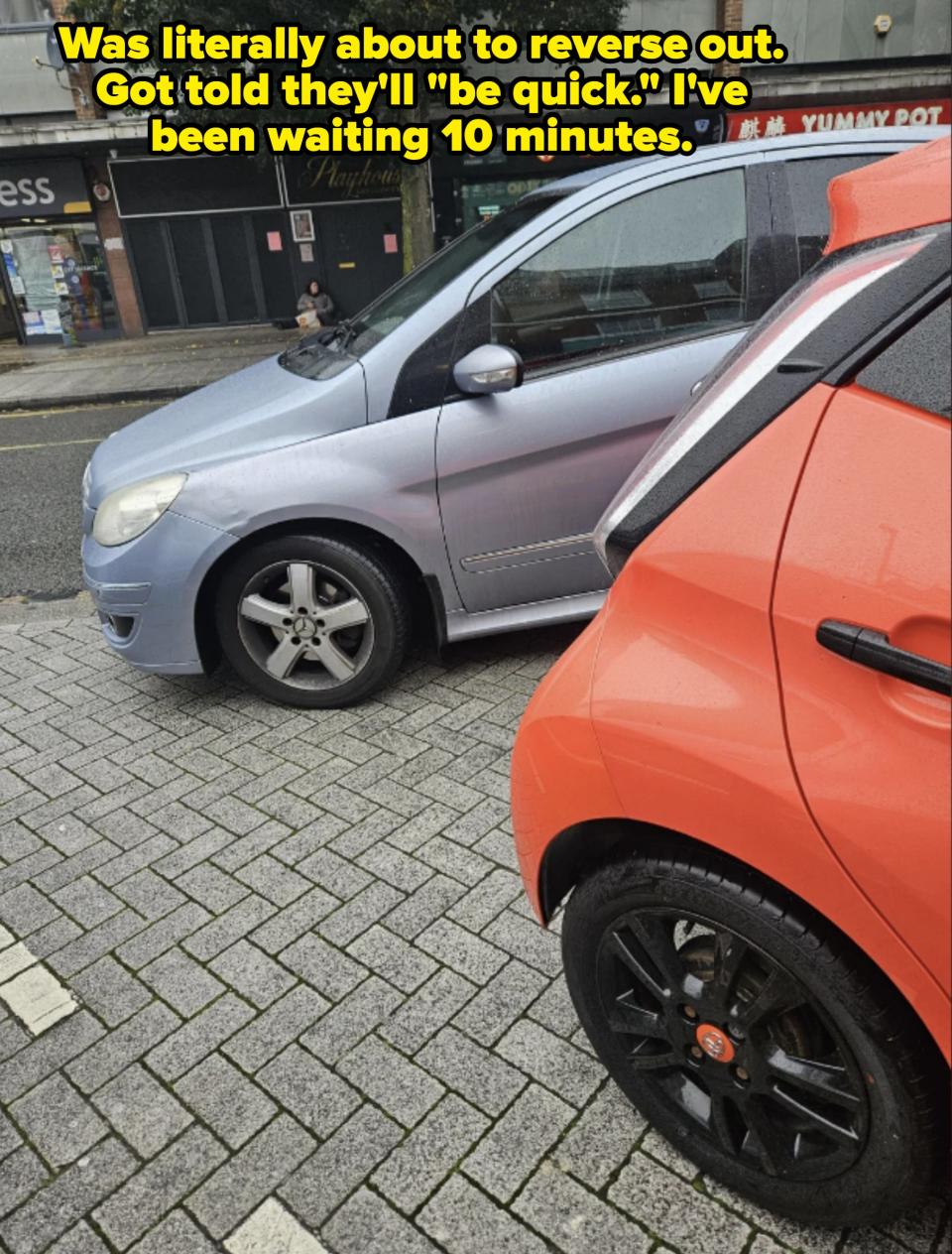 Two parked cars on a street; a blue car in front and an orange car behind. Visible storefronts and restaurants in the background