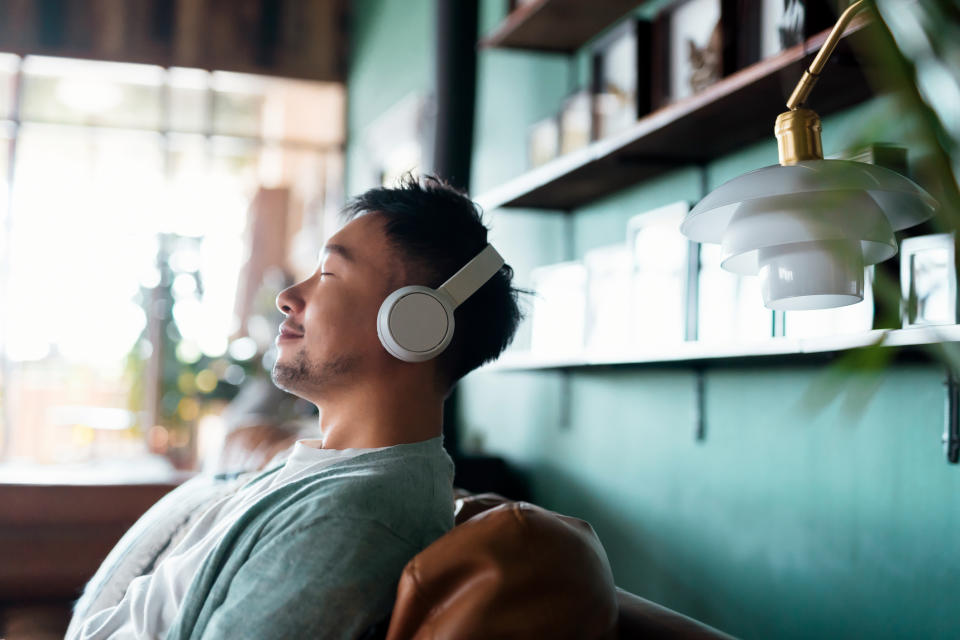 A man sits on a leather chair wearing headphones, eyes closed, seeming relaxed in a modern home setting with shelves and a hanging lamp in the background