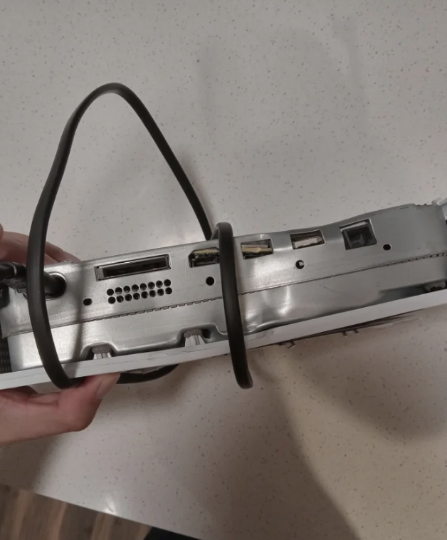 A hand holds the back of an old gaming console with various ports, cords, and connectors visible