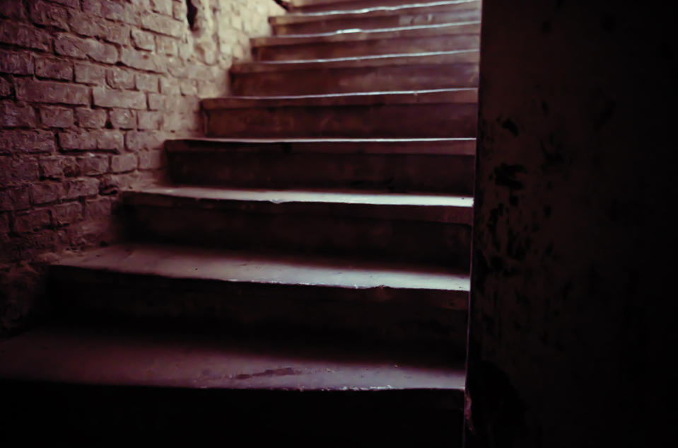 Dimly lit concrete staircase within a rustic, exposed brick-walled structure, leading upwards. The area appears aged and neglected