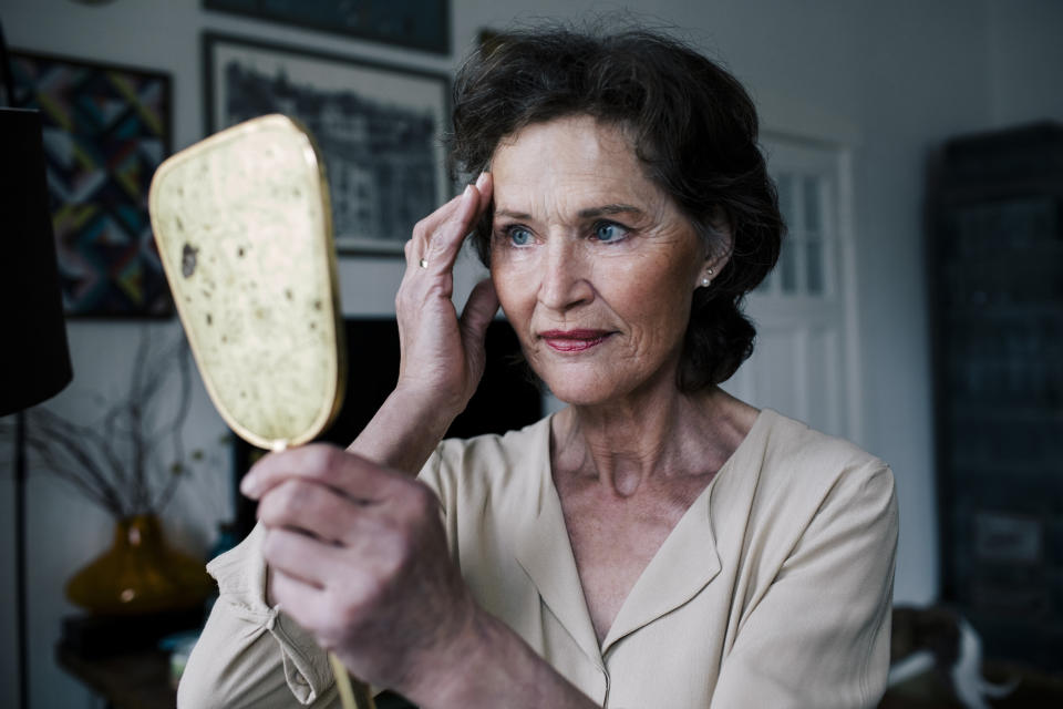 An older woman in a beige blouse examines her reflection in a handheld mirror at home