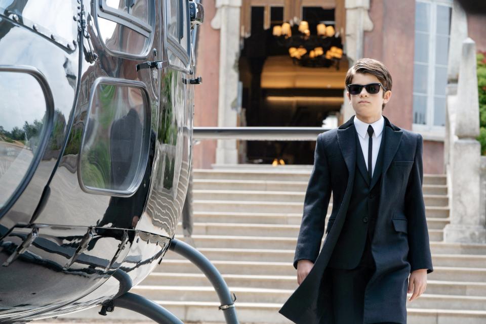 Artemis Fowl character in a suit, tie, overcoat, and sunglasses, walking confidently towards a helicopter, in front of a grand building with stairs