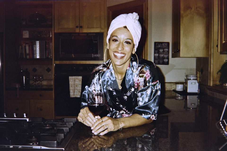 Person with a big smile, wearing a robe and headwrap, holds a glass of wine while leaning on a kitchen counter
