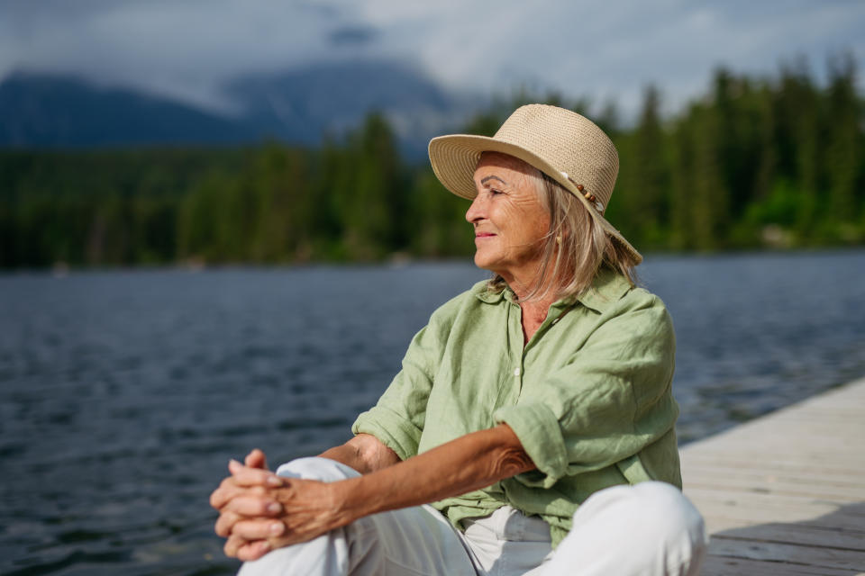 An older woman in a straw hat and relaxed green shirt sits peacefully on a dock by a forested lake, gazing into the distance with a serene expression