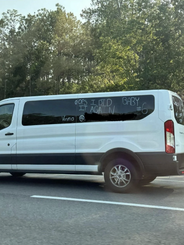 White van with "Oops I did it again, baby #6" written on the side, along with a Venmo handle, driving on a highway with trees in the background