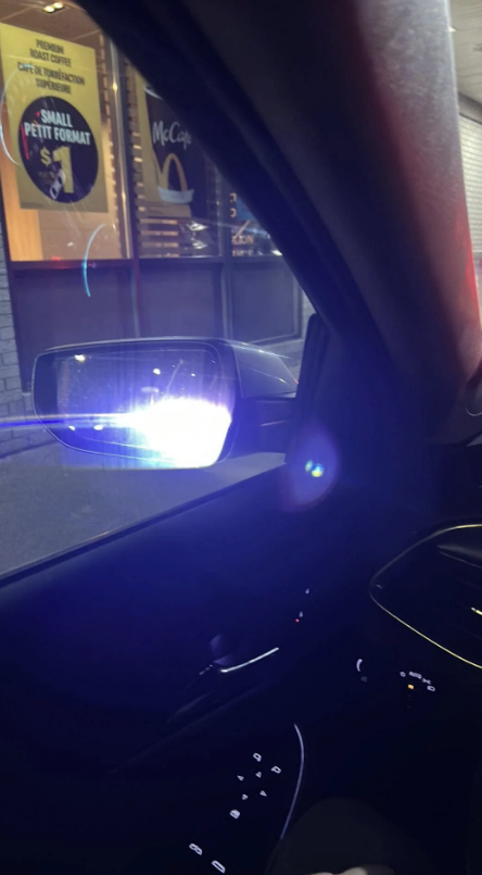 A police car is visible in the side mirror of a car, with its lights on, near a McDonald's drive-thru window
