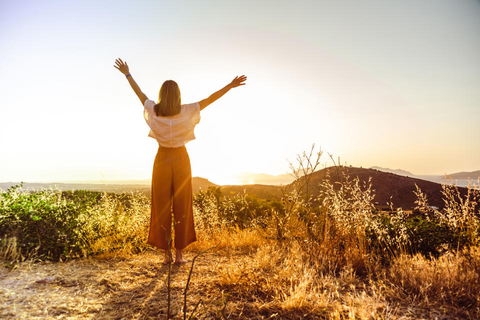 Person stands with arms outstretched, facing a sunrise or sunset over hills. The scene depicts a sense of freedom and tranquility