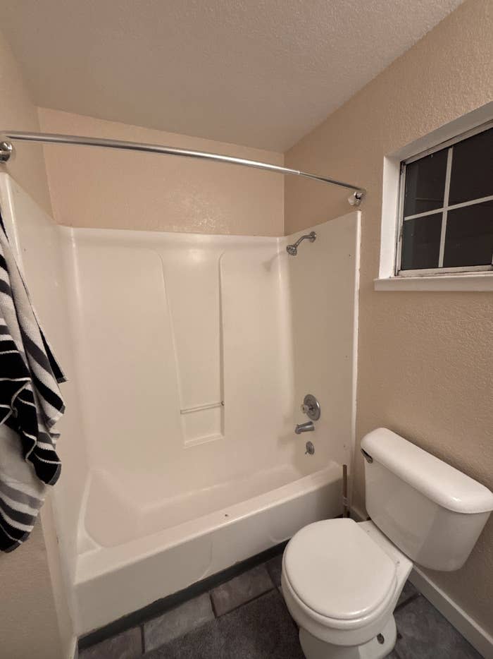Bathroom with a white shower-tub combo, a toilet, a window, and a towel hanging on a rod on the left wall
