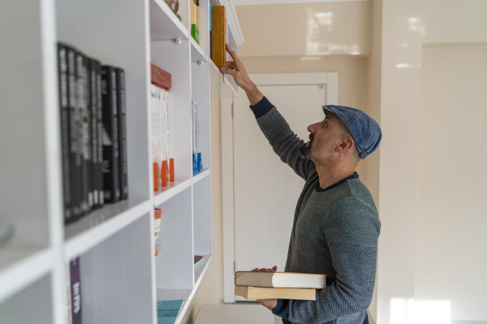 A man in a cap and sweater reaches for a book on a high shelf while holding two other books in his left hand. Shelves are filled with various books