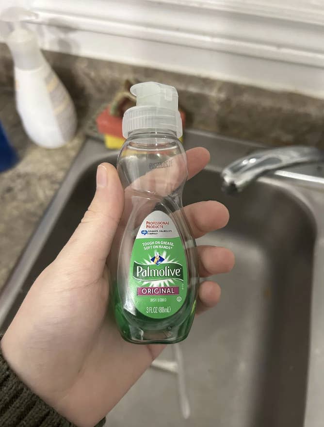 A hand is holding a small bottle of Palmolive Original dish soap above a kitchen sink