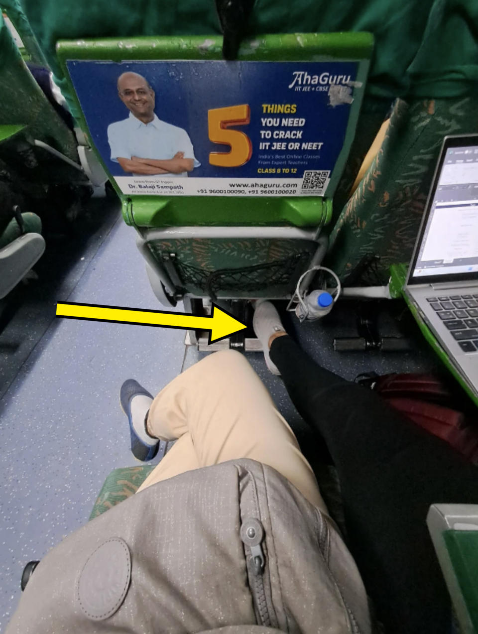 Two people seated, one in white pants, and the other in black, on a bus. An advertisement for AhaGuru with a man and text about "5 things you need to crack IIT JEE or NEET" is visible