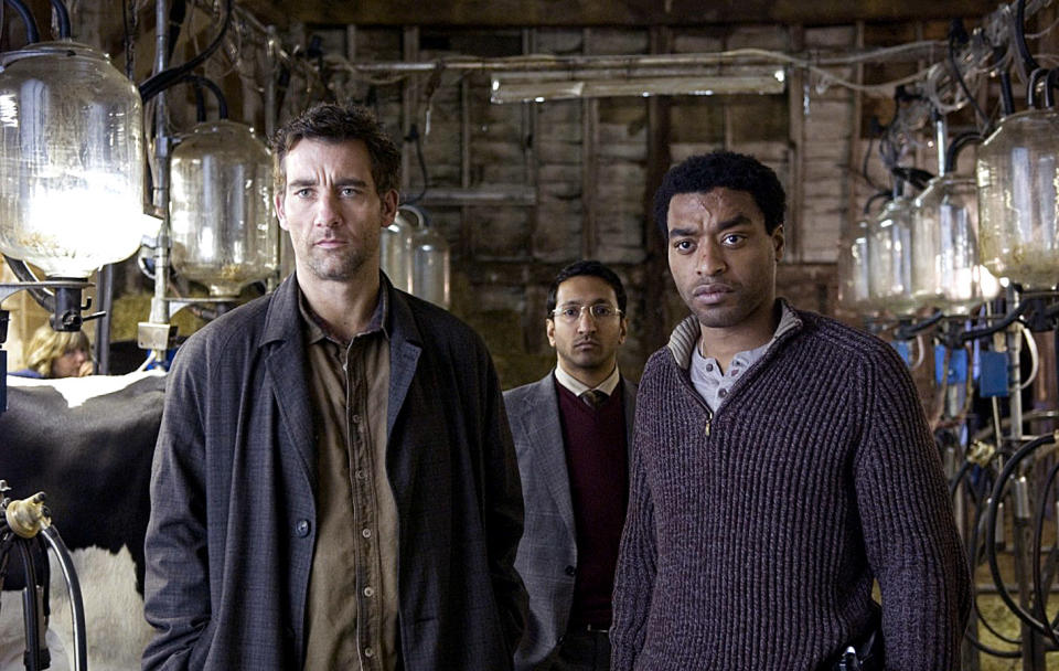 Clive Owen, Chiwetel Ejiofor, and another man stand inside a dimly lit barn with milking equipment in the background