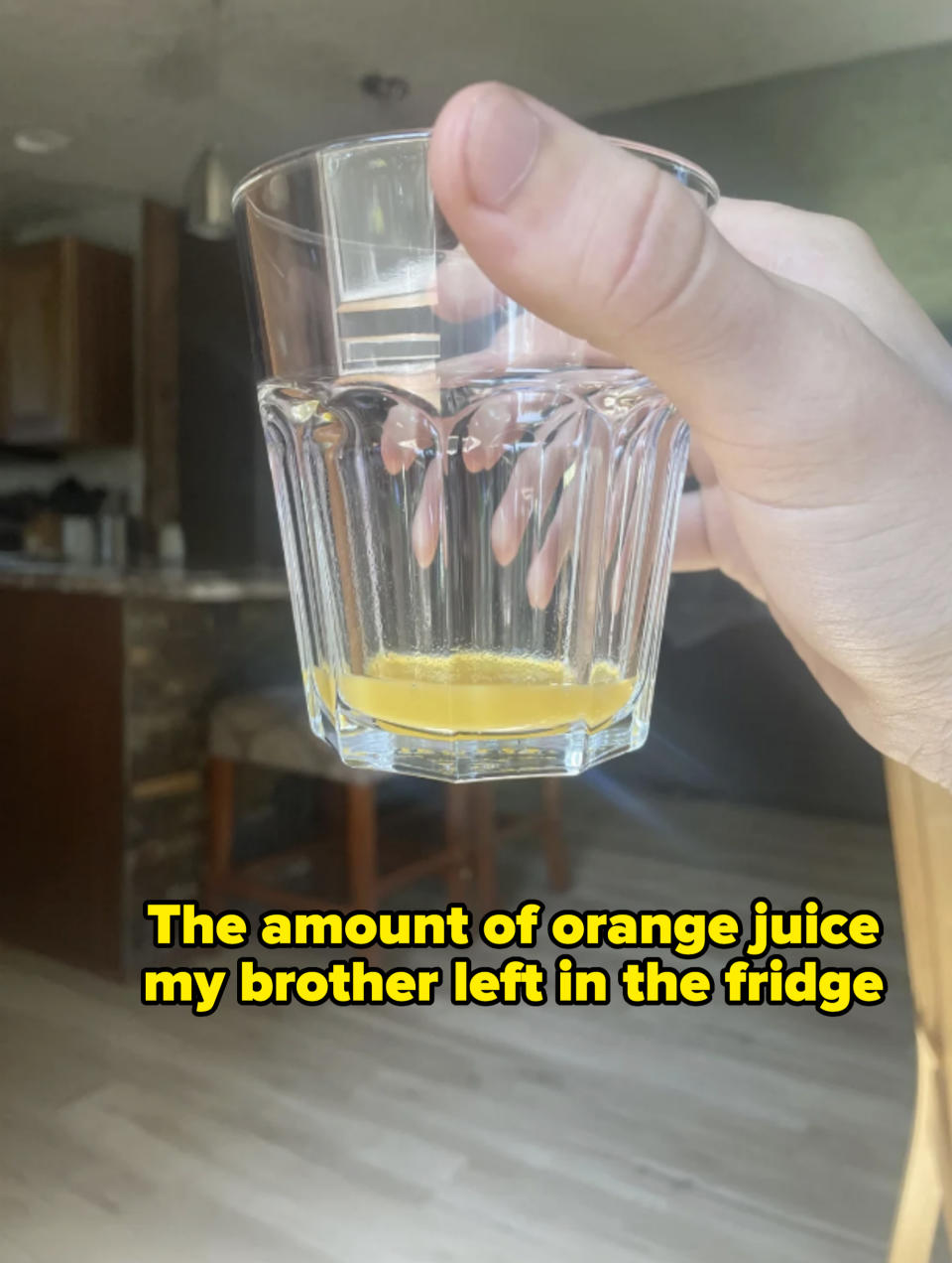 A hand holds a nearly empty glass with some yellow liquid residue at the bottom. The background shows a kitchen area with stools and wooden flooring