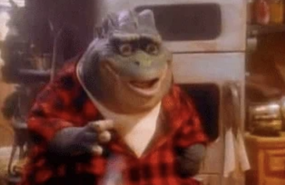 Earl Sinclair from the TV show Dinosaurs wearing a red plaid shirt, gesturing with his hand, with a backdrop of a kitchen scene