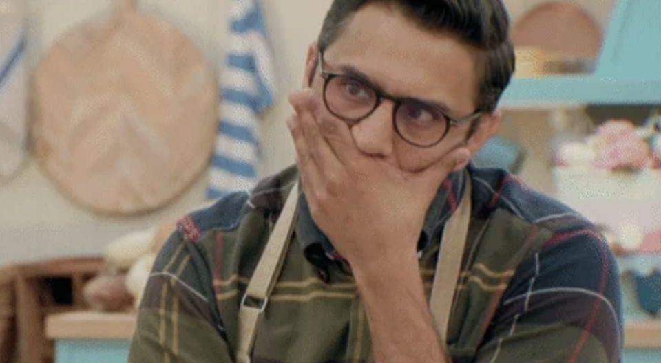 A man wearing glasses and an apron covers his mouth with his hand, appearing surprised or shocked, in a kitchen setting