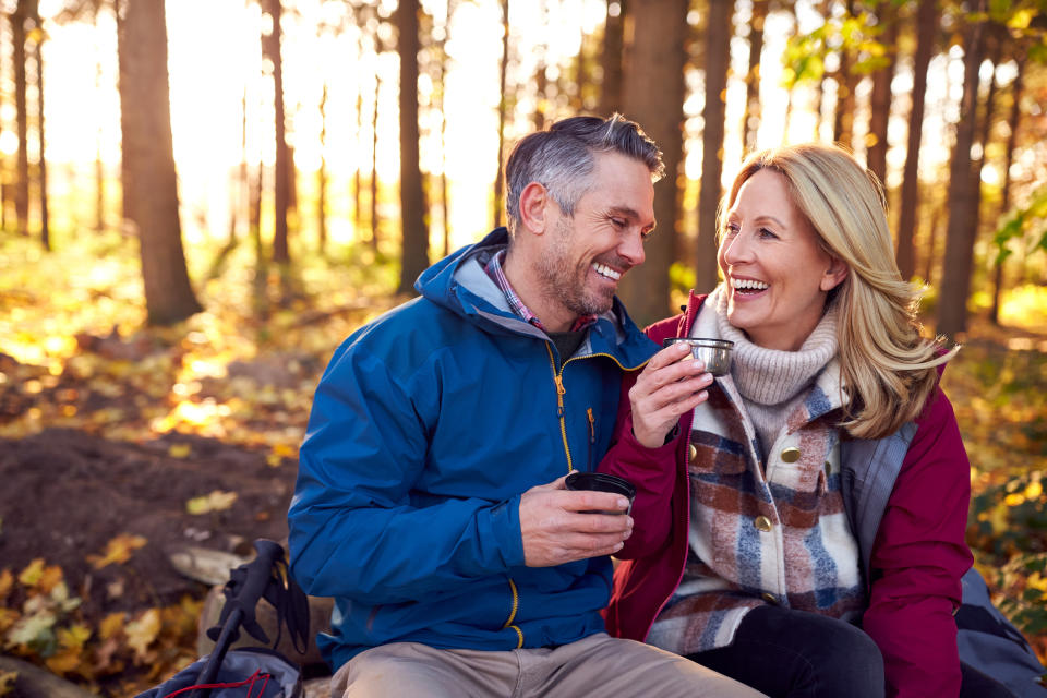 Middle-aged couple smiling and sharing a drink while sitting in a forest during autumn