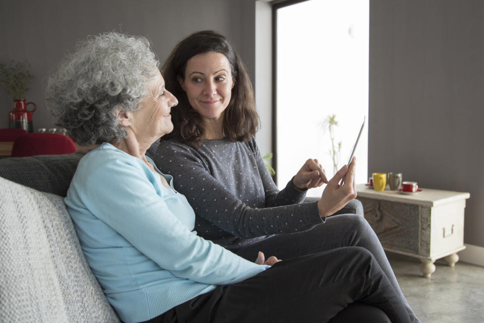 Two women, one elderly with grey hair and a light top, the other younger with dark hair and a dotted sweater, sitting on a couch smiling while looking at a tablet