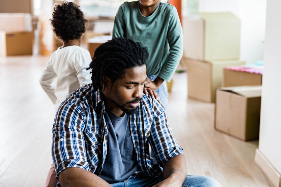 A man sits on the floor looking down, surrounded by moving boxes, with two children in the background