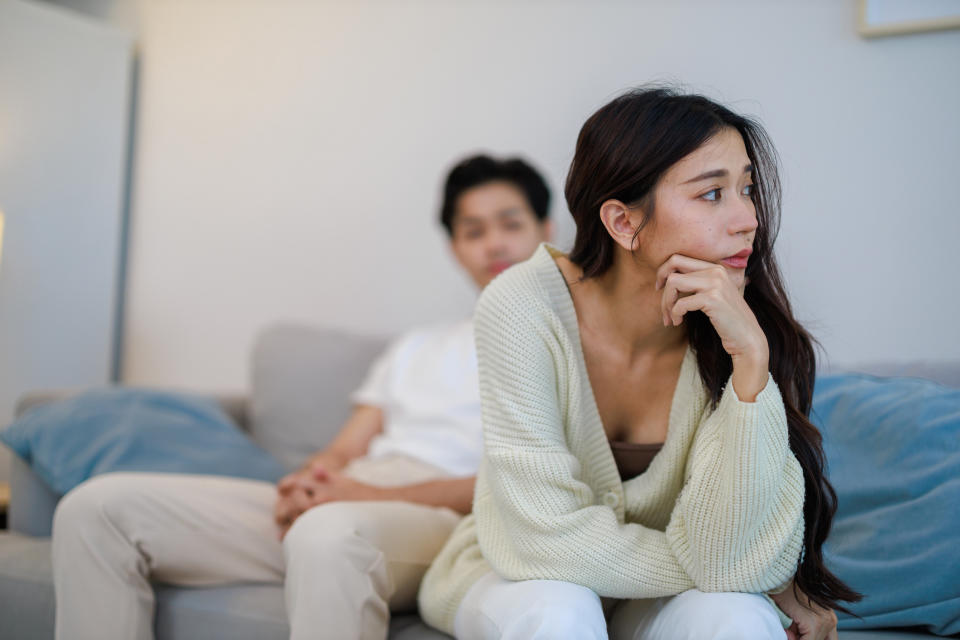 A woman with long hair sits on a couch looking pensive, with a man in the background. Both are casually dressed in comfortable clothing