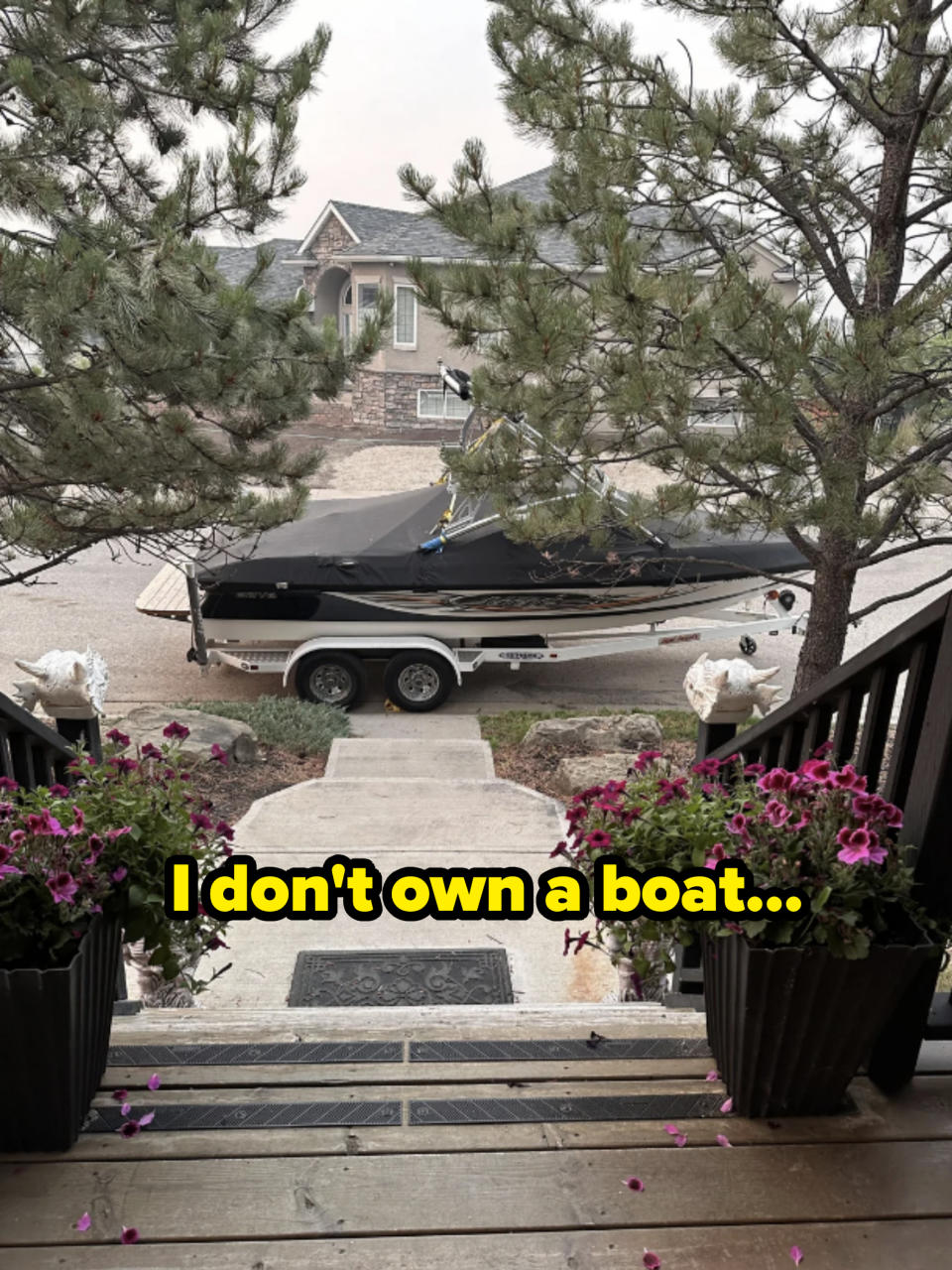 A boat on a trailer parked in a driveway, framed by trees and flowers in pots on either side of the steps leading to the house in the background