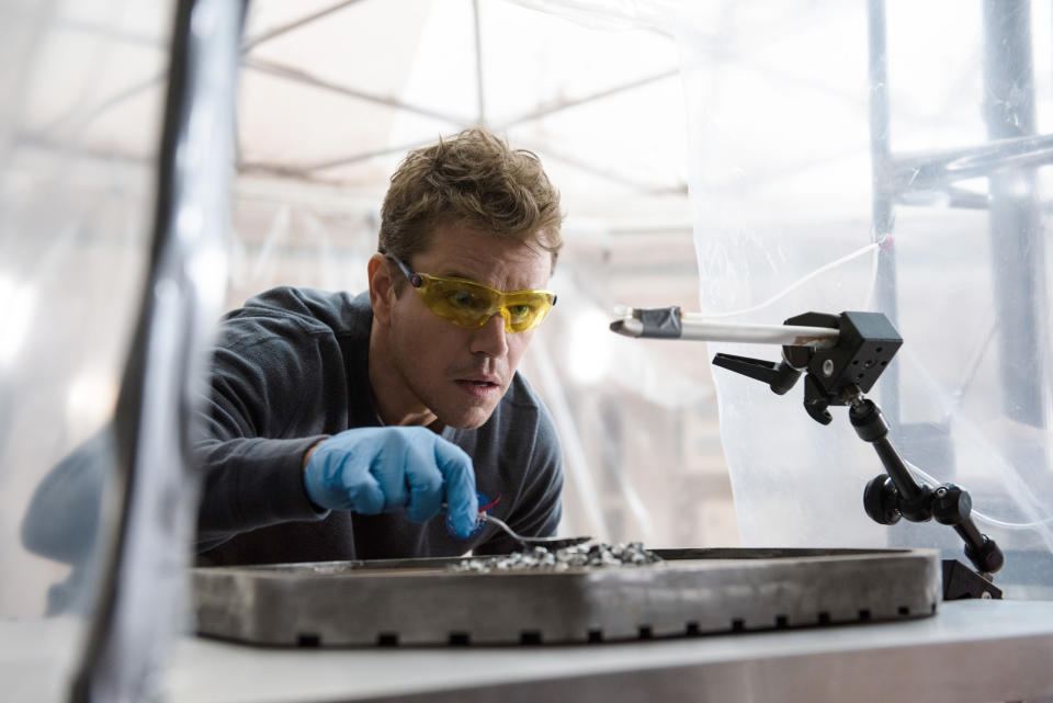 Matt Damon in a laboratory setting, wearing safety goggles and gloves, examining materials with a robotic arm
